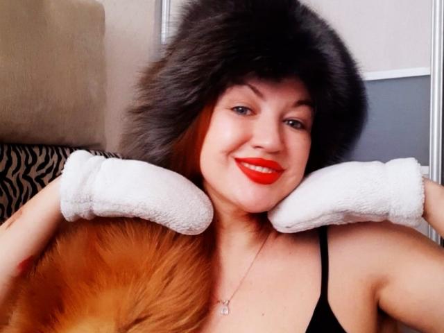 KiraXTender with red lips, black fur hat, white fuzzy cuffs, black tank top, and orange hair, smiling at camera. Her tits are visible, and she's wearing a necklace.