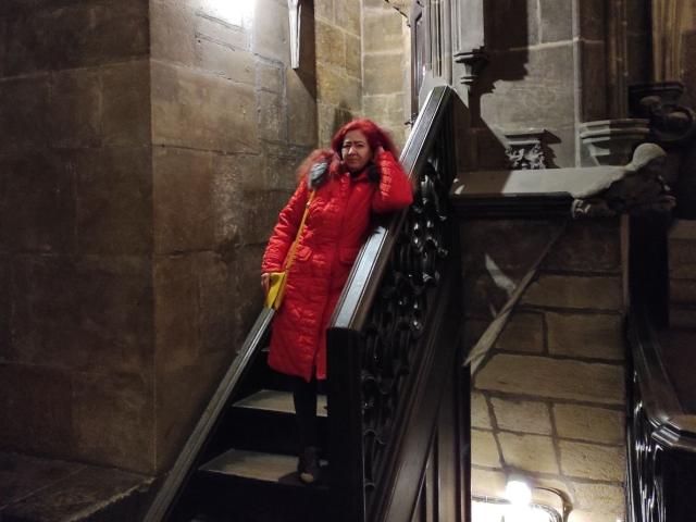MilayaDFlow se tient sur un escalier sombre, vêtue d'un manteau rouge matelassé, d'un pantalon noir et de lunettes, avec un sac jaune, et tient sa tête dans ses mains dans une zone faiblement éclairée, aux murs en pierre..
