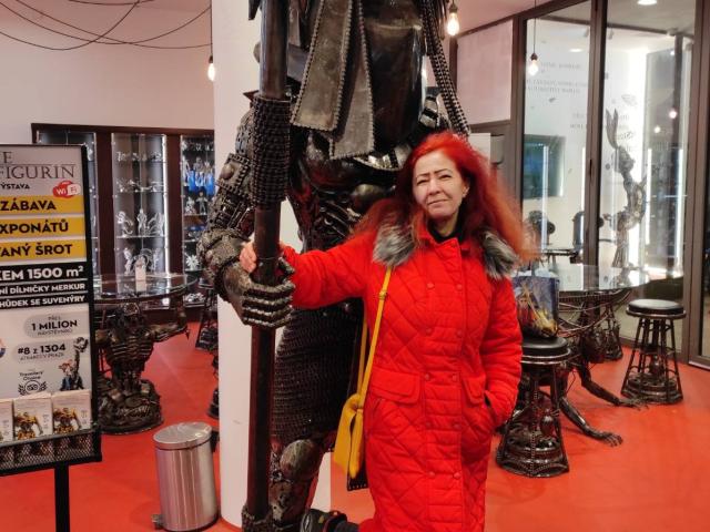 MilayaDFlow mit leuchtend roten Haaren und einem roten, gesteppten Mantel, steht neben einer großen Metallstatue mit strukturierter Rüstung und hält deren Hand. Ein gelber Riemen liegt über ihrer Schulter..