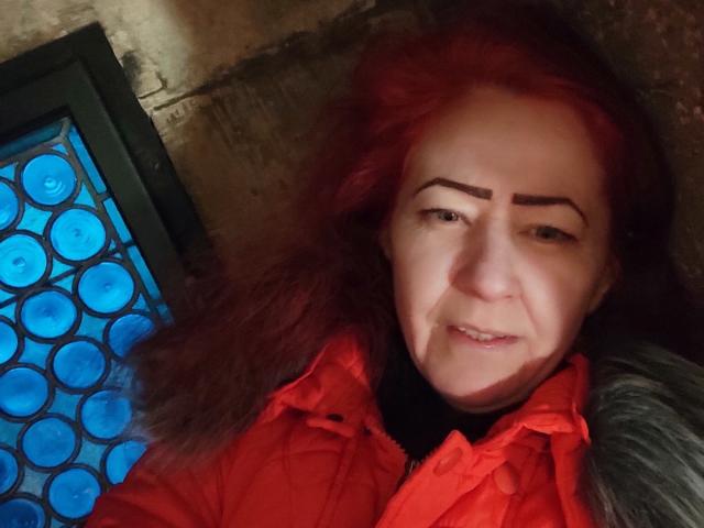 MilayaDFlow, avec des cheveux roux, des sourcils arqués et un léger sourire, portant une veste rouge, allongée avec une fenêtre en verre bleu sur la gauche..