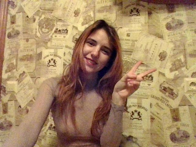 RoseJill, com longos cabelos castanhos, sorrindo e fazendo um sinal de paz, sem blusa, mostrando pequenos seios, em um quarto com pouca luz, com um papel de parede com o padrão de mapas antigos..