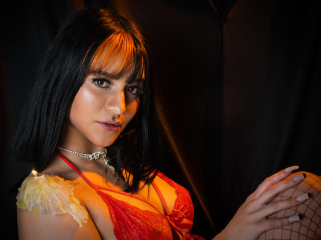 LindaWatsong con un corte de pelo bob negro, un sujetador de encaje rojo, una flor amarilla, medias de red, un piercing en la nariz y un collar, irradiando en una luz naranja, sosteniendo su rodilla, mirando seductoramente a la cámara..