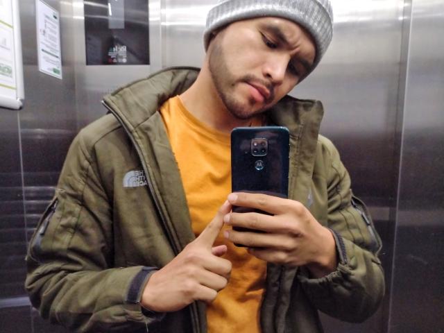 Maotroz, com um gorro cinza, jaqueta verde e camisa laranja, aponta para a câmera do celular, tirando uma selfie em um elevador de metal. Seu rosto mostra um leve semblante de descontentamento..
