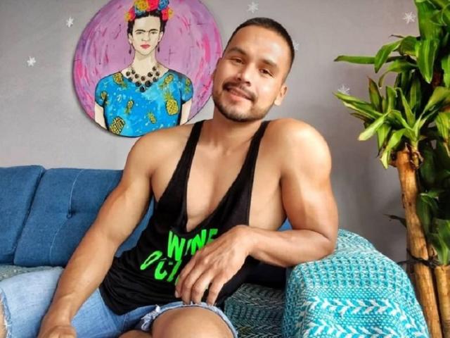 Maotroz with a muscular build, tan skin, short hair, and beard, wearing a black tank top showing his sideboob, and blue jeans, sitting on a couch with a green plant.