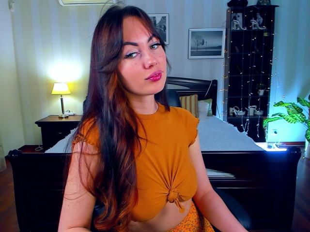 NinaTender with long brown hair, orange crop top, tied below bust, showing small tits, sitting in bedroom, pink lipstick, subtle makeup, wooden bed, lamp, shelf, plant in background.