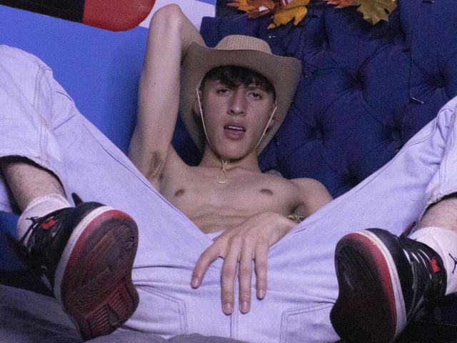 Carlos Rubiio, sem camisa, usando um chapéu de cowboy, calças brancas e tênis pretos com solas vermelhas, pernas abertas, mão na virilha, sofá azul com textura, folhas de outono ao fundo..