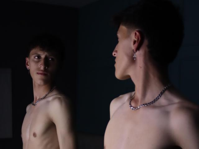 Carlos Rubiio è senza camicia, indossa una collana e un orecchino, ha i capelli corti e scuri, ed è in piedi in una luce fioca, guardando il suo riflesso in uno specchio. Il suo petto è visibile..