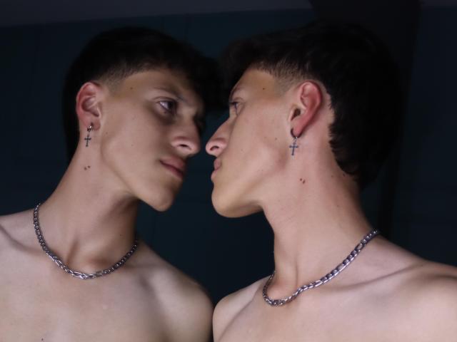 CarlosRubiio, shirtless with cross earrings and chain necklaces, stares into mirror reflection, showing smooth pale skin, short dark hair, and serious expressions. Mirrored faces, intense eye contact.