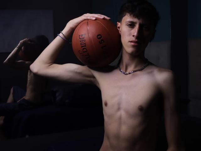 Carlos Rubiio, sem camisa, com um corpo magro e tonificado, segurando uma bola de basquete Spalding no ombro direito, usando um colar de corrente, em um quarto com pouca luz e um espelho..