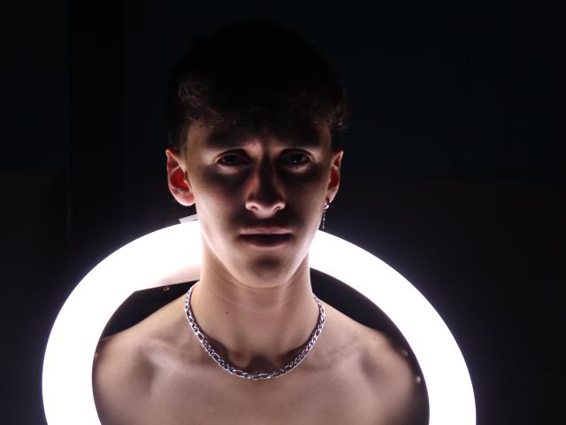 Carlos Rubiio, zonder shirt, met een zilveren ketting, staat voor een heldere ringlamp, donkere achtergrond, kort bruin haar, serieuze uitdrukking, lichte schaduw op zijn gezicht..