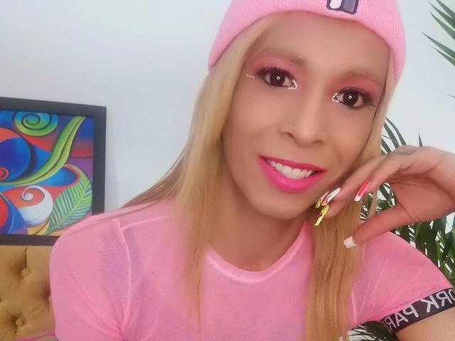 FlaquitaRaquel con capelli biondi, berretto rosa, ombretto rosa, rossetto rosa, maglietta rosa e unghie lunghe, sorridente, con un piercing al naso, la mano destra che tocca il mento, con opere d'arte colorate sullo sfondo..