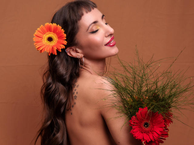 ElizaWells with long brown hair, orange flower in hair, red lipstick, hoop earrings, star tattoo on back, holding grass and red flower, smiling, topless, brown background.