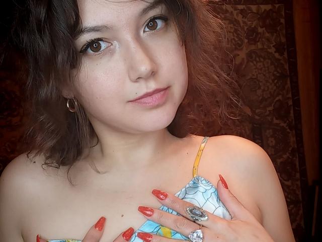 CarriePeachy, con cabello castaño ondulado, vestida con un vestido colorido, cubriendo sus senos con manos de uñas rojas, ojos marrones, nariz pequeña, sonrisa sutil y un anillo grande en su mano derecha..