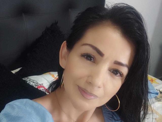 YourDulceHott69 tem pele morena, cabelo preto, olhos castanhos, usa brincos de argola dourados, tem um sorriso discreto e lábios rosados, veste uma blusa azul e está olhando para a câmera..