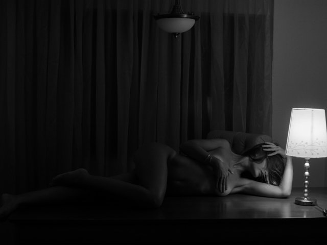 MarCrissa liegt nackt auf einem Tisch, ihre Brüste und ihr Hintern sind teilweise sichtbar, unter einer Lampe, mit der Hand auf dem Kopf, in einem dunklen, stimmungsvollen Foto..