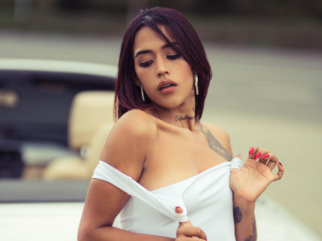 LiaClarck, avec ses cheveux roux foncés, un haut blanc à épaules dénudées, laissant entrevoir ses seins, qu'elle retient, des ongles roses, des tatouages, un collier ras du cou, le regard baissé, une voiture en arrière-plan, une lumière douce..