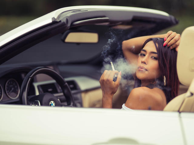 LiaClarck with dark brown hair, smoking a cigarette, leaning in a white BMW convertible, hand on head, looking at camera, red nails, beige interior, relaxed expression.