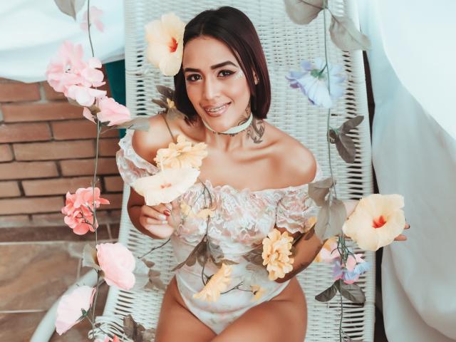 LiaClarck with brown hair, smiling, wearing lace off-shoulder lingerie, sitting on white wicker chair, surrounded by pink and peach flowers, showing her tits, with a choker.