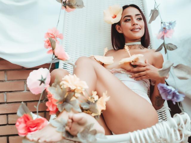 LiaClarck with dark hair, wearing a white lace top, sitting in a white wicker chair, surrounded by pink and purple flowers, holding petals, smiling, showing her tits and ass.