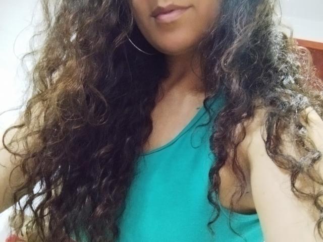 Angelonix with long, curly brown hair wearing a green tank top, showing her small tits and cute face, wearing large hoop earrings, slightly smiling, in a casual selfie.
