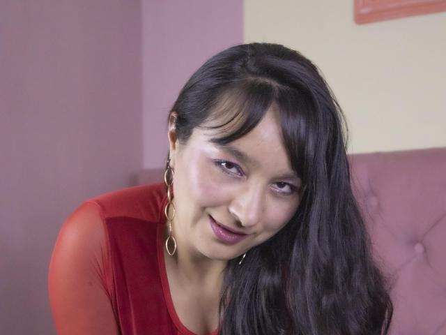 LexiLuna, avec ses longs cheveux noirs, portant un haut rouge transparent, de grandes boucles d'or, souriant légèrement et regardant l'appareil photo, mettant en valeur son joli visage et son maquillage discret..
