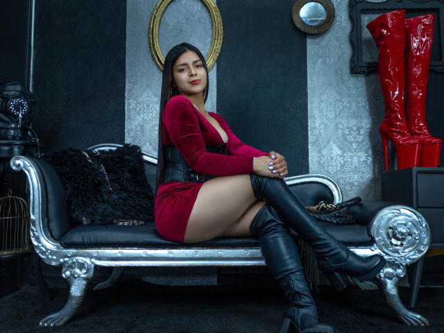 Demi Stuart, con cabello negro largo, vestido de terciopelo rojo, corpiño negro y botas negras hasta la rodilla, sentada en un sofá plateado ornamentado, mostrando su trasero y luciendo segura de sí misma..
