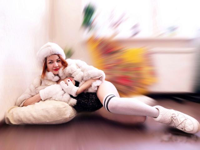 KiraXTender con cabello rojo, un sombrero de piel blanca, un abrigo esponjoso, un vestido de encaje negro, calcetines blancos y zapatillas blancas, acostada sobre un cojín, sosteniendo un peluche blanco. Fondo colorido y borroso..