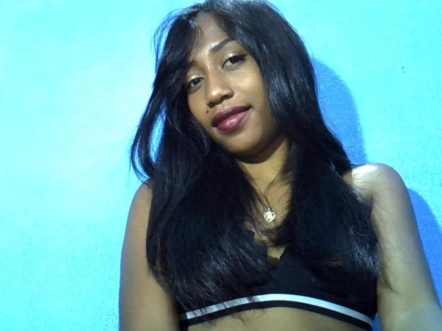 A AmaraCute tem cabelos pretos longos, pele morena e está vestindo um top esportivo preto com listras brancas, olhando ligeiramente para o lado, com uma pequena pinta na bochecha direita..