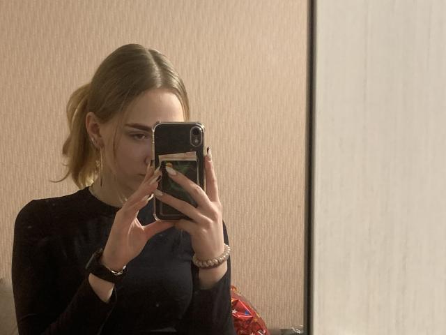 Une jeune femme blonde avec des seins de taille moyenne, un haut noir, une queue de cheval blonde, des boucles d'oreilles dorées, un bracelet noir, tenant un téléphone, avec une expression sérieuse, prenant un selfie dans un miroir..