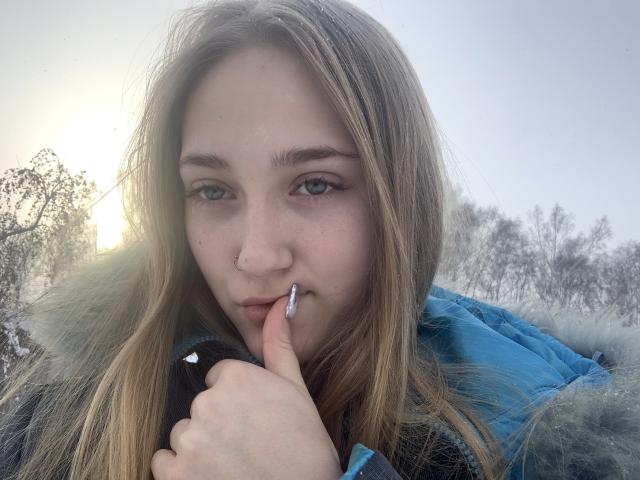 Garota loira com olhos azuis, pele clara e cabelo loiro, mordendo o dedo, vestindo uma jaqueta azul acolchoada com detalhes de pele, em um ambiente externo com neve..