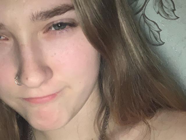 A GoodBlondeGirl tem pele clara, olhos castanhos e um piercing no nariz. Ela está sorrindo levemente, com cabelos castanhos longos e um leve rubor nas bochechas. Seus seios não estão visíveis..