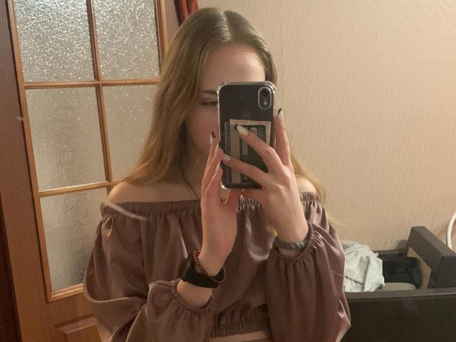 Blond meisje met lang, blond haar, een roze-bruine top met open schouder, die een telefoon voor haar gezicht houdt, spiegelzelfportret, draagt een zwarte armband..