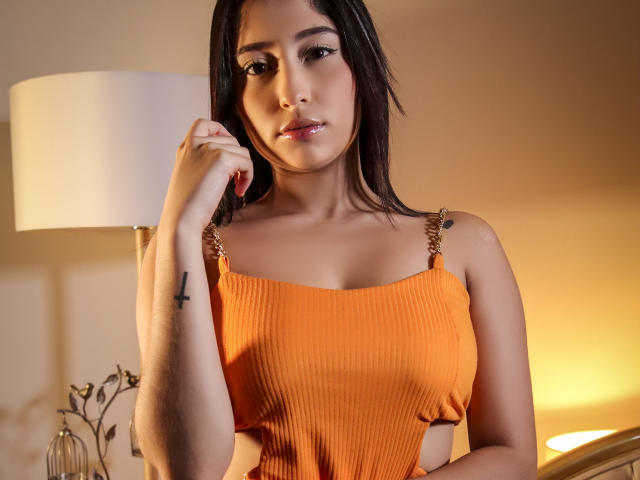 ArianaCandyX with medium tits in an orange ribbed top, dark hair, and a tattoo on her left arm, looking slightly down with a hand near her face.