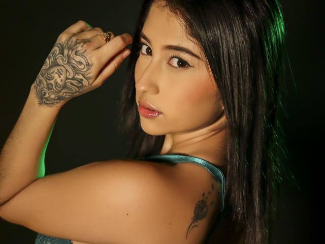 ArianaCandyX, con cabello oscuro, ojos marrones, labios rosados brillantes, tatuaje en la mano derecha, tatuaje en el hombro izquierdo, vistiendo una blusa verde turquesa, mirando por encima del hombro, luz verde..