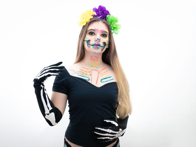 PaolaYKatty, con maquillaje de calavera de colores en la cara, una blusa negra, guantes de esqueleto, una diadema de flores y cabello castaño largo. Sus senos son visibles y está sosteniendo un guante..