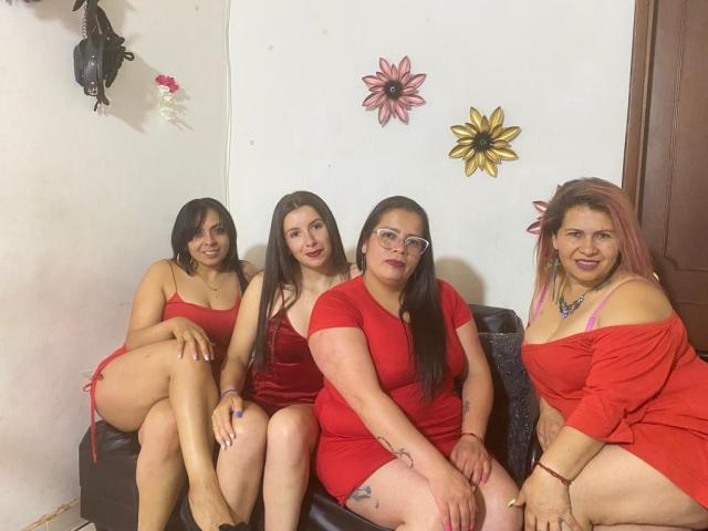 Vier lesbische Frauen mit langen, dunklen Haaren, roten Kleidern und Brillen sitzen auf einer schwarzen Couch. Zwei haben kleine Brüste, eine trägt eine Brille, eine hat pinke Haare. Hintern und Tattoos sind sichtbar..