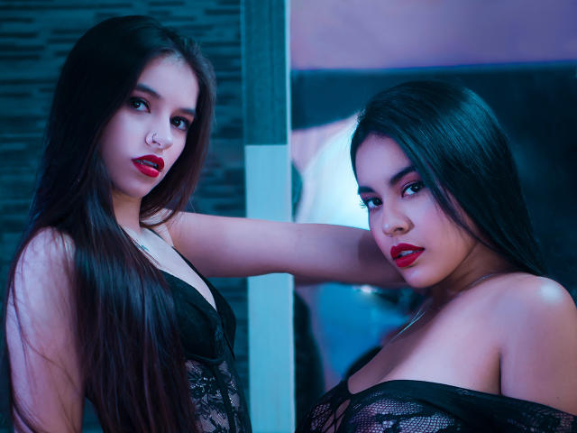 PaolaYKatty with long dark hair, red lipstick, nose piercing, and black lace lingerie, standing in front of a mirror, showing her tits and ass reflection, in blue lighting.