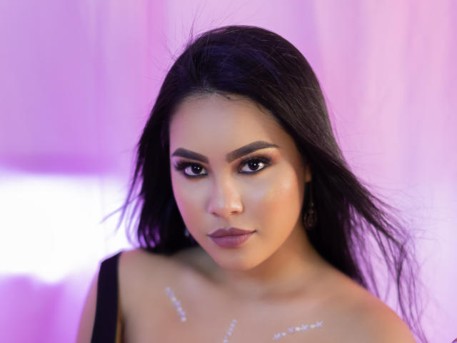 PaolaYKatty has dark, straight hair, brown eyes, and is wearing sparkly makeup with glitter on her chest, looking forward with a slight smile, in a pink-lit photo.