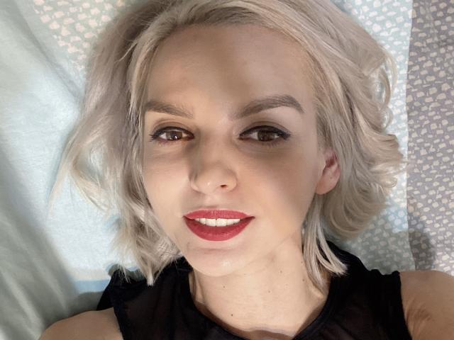 DouceAmelia has platinum blonde hair, brown eyes, red lipstick, and black eyeliner. She's smiling with a cute face, wearing a black top, lying down.