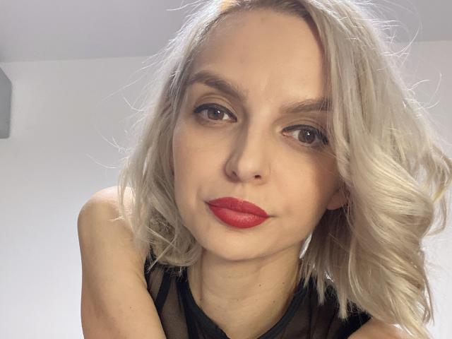 DouceAmelia with platinum blonde hair, red lipstick, and black sheer top, looking directly at the camera with a slightly pouty expression. Her tits are partially visible.