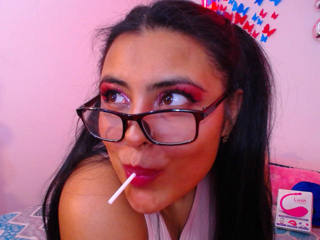 AngelaRoggers with black hair, wearing glasses, pink eyeshadow, red lipstick, and a lollipop in her mouth. Her tits are slightly visible. Pink wall in background.