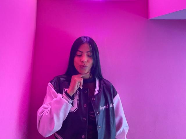 Amberv with long black hair, wearing a black and white varsity jacket, touching her chin with a subtle smile, bathed in pink and purple neon light.