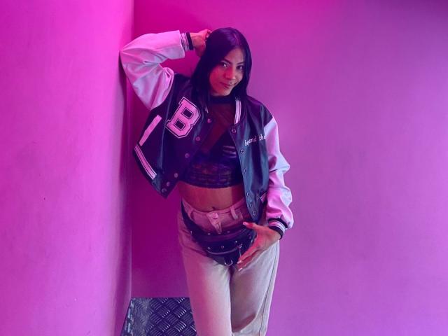 Amberv with long black hair, wearing a black lace bra, gray varsity jacket, and beige pants, stands against a pink wall, hand on hip, smiling at the camera.