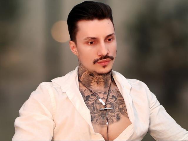 TyagoDoll, mit Tattoos, trägt ein offenes weißes Hemd, das Brusttattoos und eine Kreuzkette zeigt. Er hat dunkles Haar, einen Schnurrbart und einen ernsten Gesichtsausdruck..