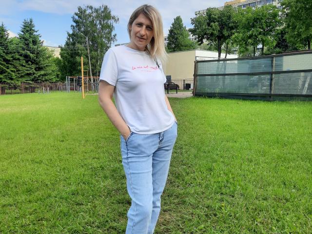 AliceShinyS steht auf einem grünen Rasen, trägt ein weißes T-Shirt, das ihre Brüste zeigt, und blaue Jeans, hat die Hände in den Taschen, lächelt und hat blondes Haar. Im Hintergrund sind grüne Bäume zu sehen..
