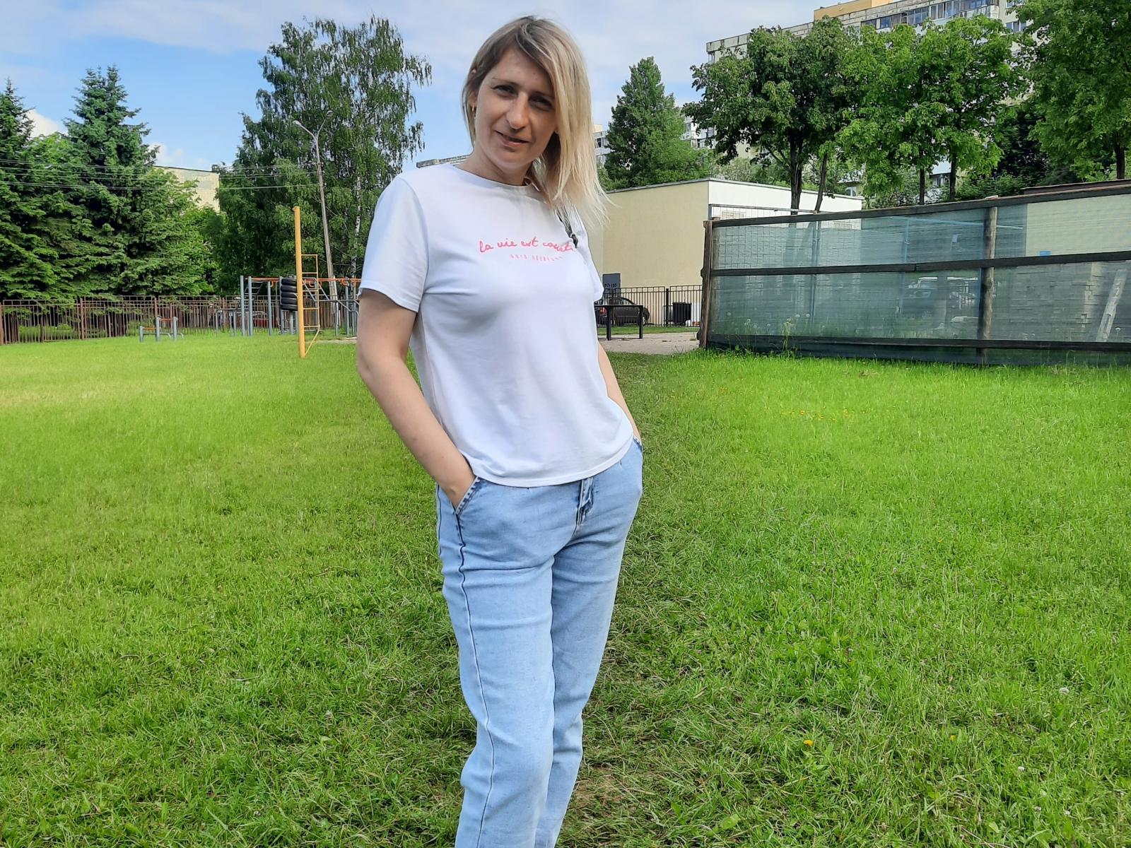AliceShinyS steht auf einem grünen Rasen, trägt ein weißes T-Shirt, das ihre Brüste zeigt, und blaue Jeans, hat die Hände in den Taschen, lächelt und hat blondes Haar. Im Hintergrund sind grüne Bäume zu sehen..