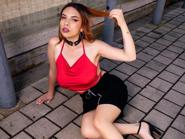 VioletaStone met een rode top met halsband, een zwarte rok en zwarte hakken, haar bruine haar vasthoudend, een zwarte choker om, zittend op een met bakstenen bestraat oppervlak, waarbij haar borsten en billen zichtbaar zijn..