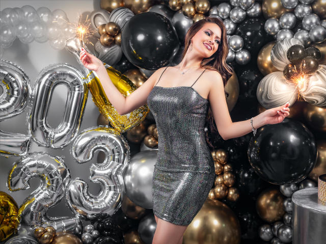 HannaRobertss in a sparkly silver dress, holding sparklers, with shiny black and gold balloons, smiling, showing off her tits and ass, with long brown hair, red lipstick.