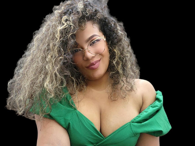 EbonyRose69 mit lockigem blondem und braunem Haar, einem grünen Kleid mit Schulterfrei-Ausschnitt, Brille und einem Lächeln, die ihre großen Brüste vor einem schwarzen Hintergrund zeigt..