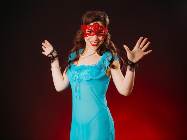 LadyXwinter mit roter Maskerade-Maske, lächelnd, trägt ein blaues Spitzenkleid, schwarze Spitzenarmstulpen und eine Perlenkette, winkt mit hochgehobenen Händen und zeigt ihre Brüste..